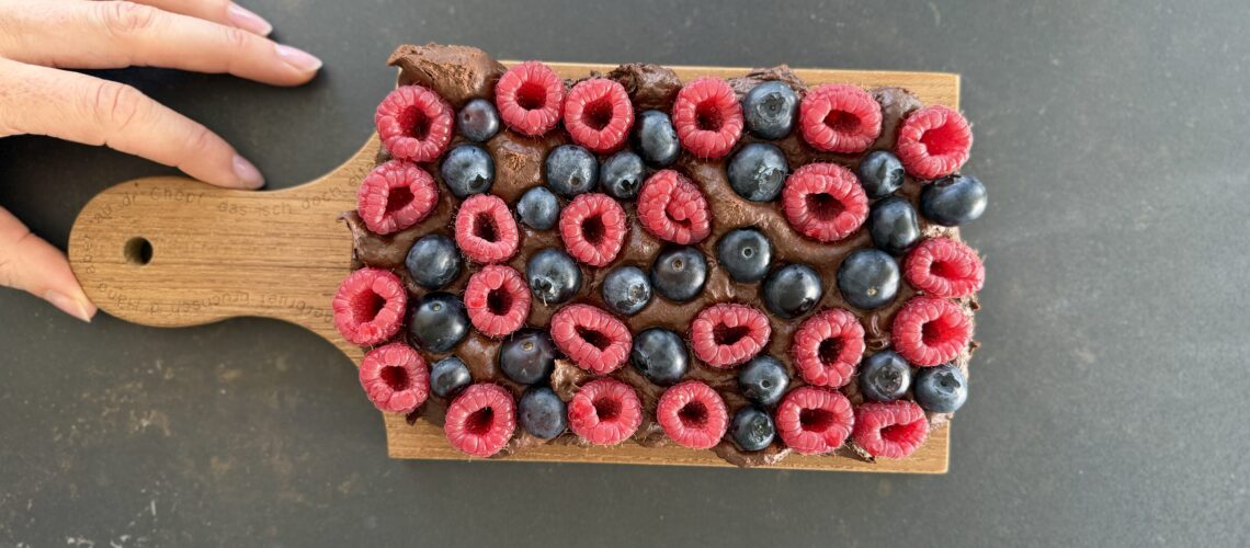Protein Fudge Gesundes Schoko Beeren Dessert auf einem Holzbrett angerichtet mit frischen Himbeeren und Heidelbeeren, cremig angeschnitten mrs flury